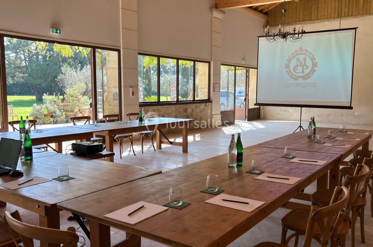 Salle de réunion lumineuse avec de grandes tables en bois, chaises, écran de projection et vue sur un jardin.