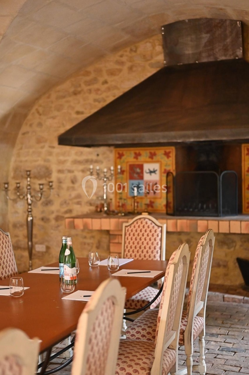 Salle en pierre avec une grande cheminée, table ronde dressée avec chaises rembourrées et bouteille d'eau.