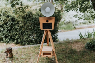 Appareil photo sur trépied en bois avec lampe circulaire, installé dans un jardin verdoyant.