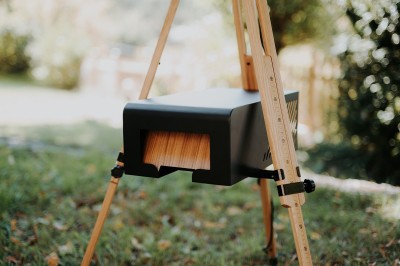 Miniature Lively Booth #3 Appareil photo en bois monté sur un trépied, installé dans un jardin avec des arbres en arrière-plan.