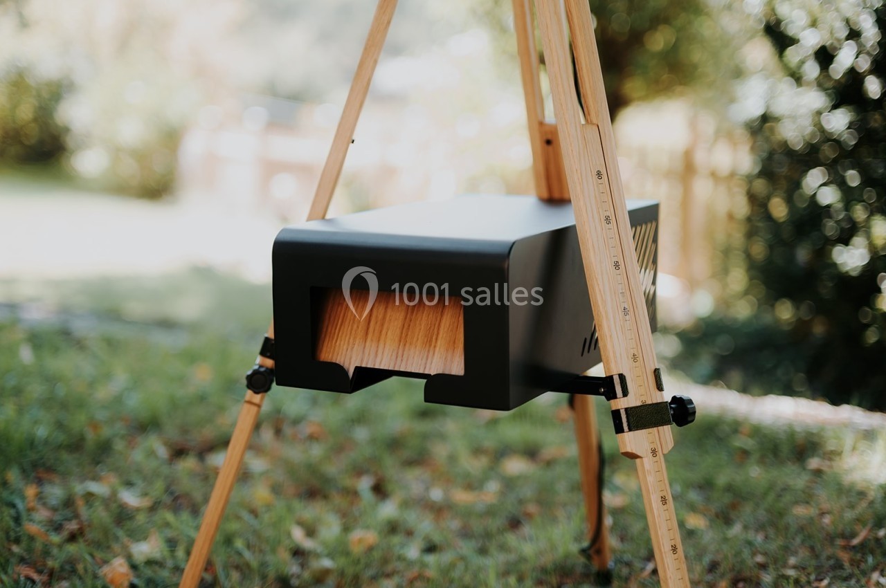 Four extérieur noir en bois monté sur un trépied en bois, placé sur une pelouse avec un jardin en arrière-plan flou.