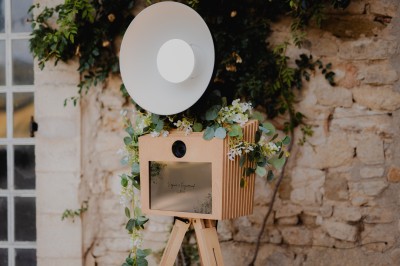 Photobooth en bois décoré de feuillage, avec une lampe circulaire, installé en extérieur lors d'un événement.