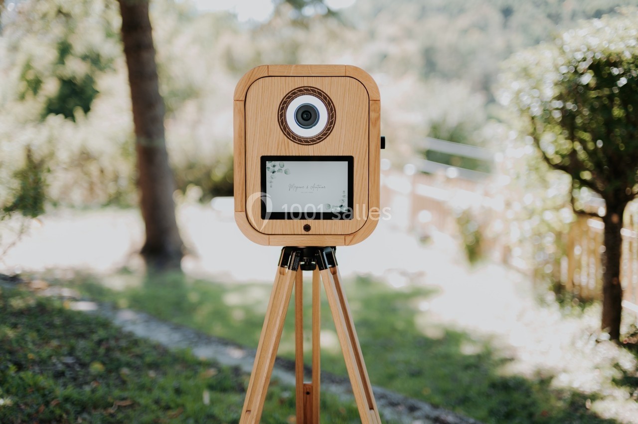 Borne photo en bois sur trépied, installée en extérieur avec un arrière-plan flou de végétation et de lumière naturelle.
