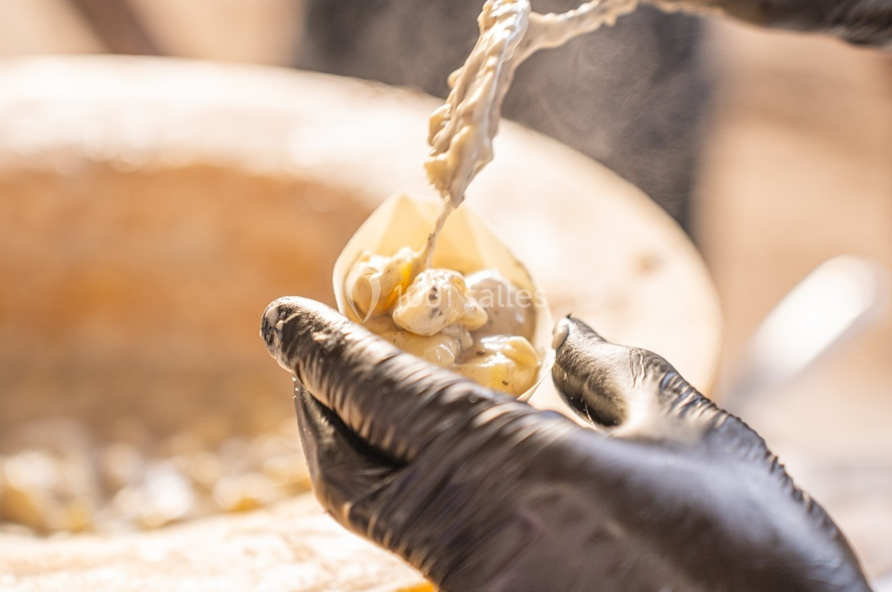 Des mains gantées servent des pâtes crémeuses dans un récipient, près d'une meule de fromage.