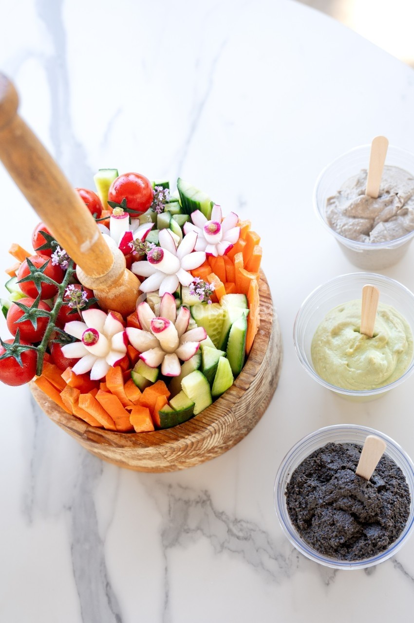 Plateau de crudités colorées avec carottes, concombres et radis, accompagné de trois sauces dans des pots séparés.
