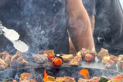 Miniature Traiteur Saint-Vallier-de-Thiey (Alpes-Maritimes) - Traiteur D'azur #13 Un cuisinier en tenue noire prépare des légumes dans une poêle en extérieur, devant un mur en pierre.