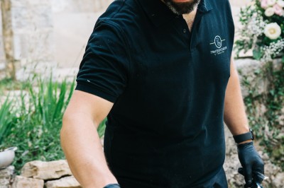 Miniature Traiteur Saint-Vallier-de-Thiey (Alpes-Maritimes) - Traiteur D'azur #14 Un cuisinier en tenue noire prépare des légumes dans une poêle en extérieur, devant un mur en pierre.