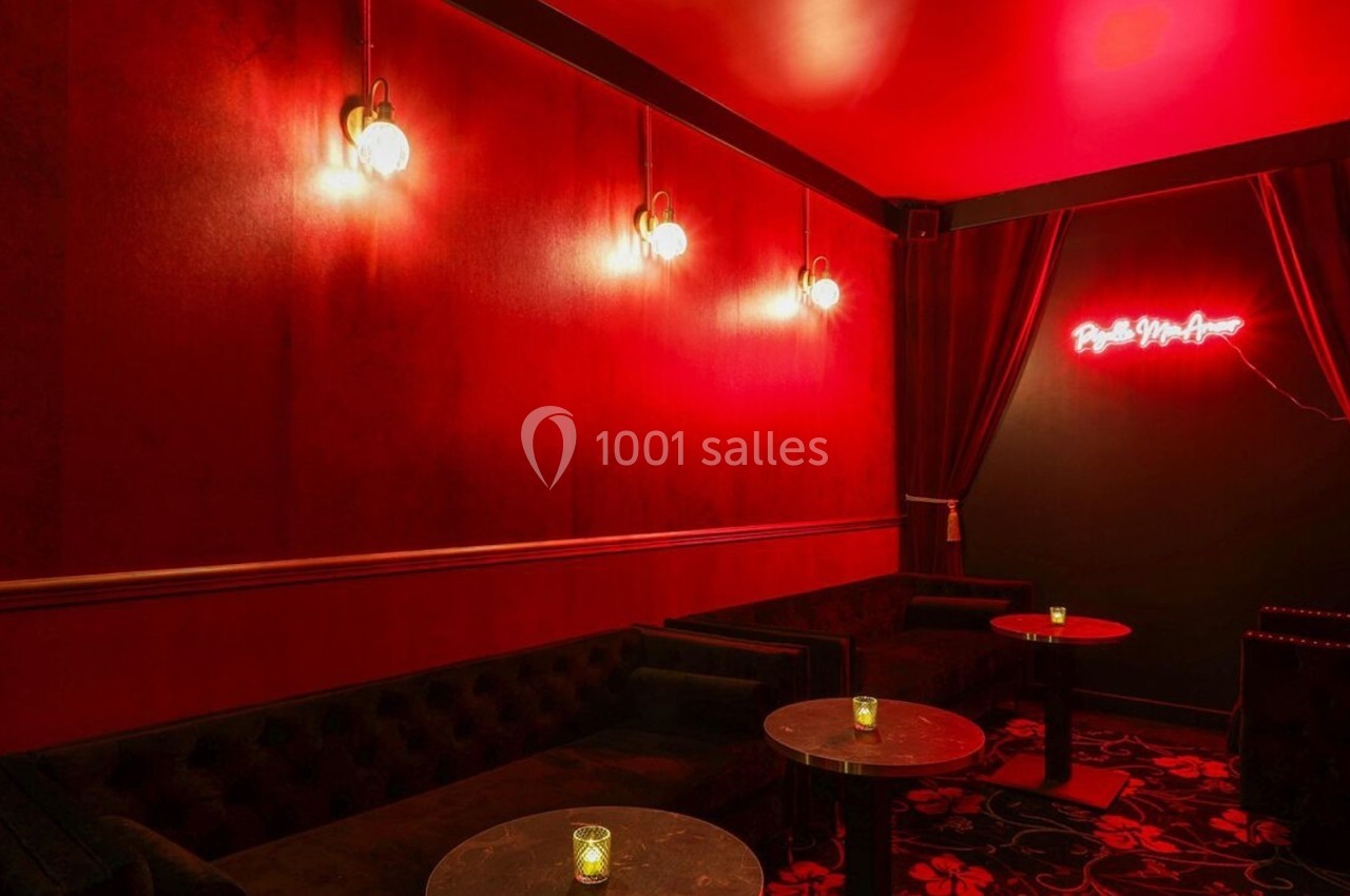 Salle au décor rouge tamisé avec banquettes sombres, tables rondes et enseigne néon lumineuse sur un mur noir.