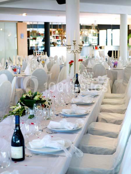 Salle de réception décorée avec des tables dressées, nappes blanches, chandeliers et bouteilles de vin.