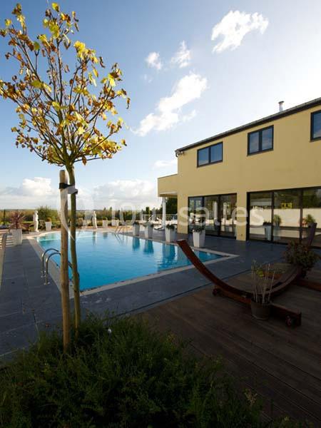 Piscine extérieure entourée de terrasses en bois et de chaises longues, devant une maison moderne sous un ciel dégagé.