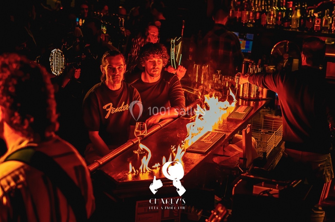Des clients assis au bar regardent un comptoir enflammé dans une ambiance de bar animée et tamisée.