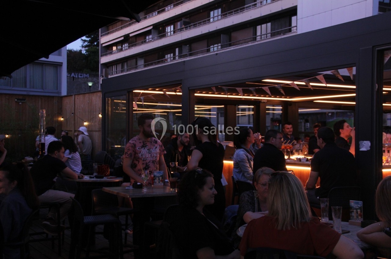 Terrasse animée en soirée avec des groupes de personnes discutant autour de tables et d'un bar éclairé.