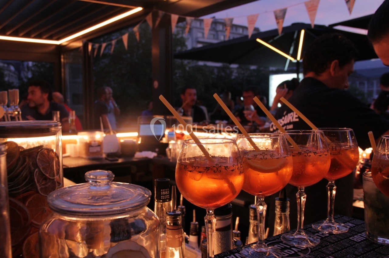 Verres de cocktails alignés sur un bar éclairé, avec des personnes discutant en arrière-plan dans une ambiance nocturne.