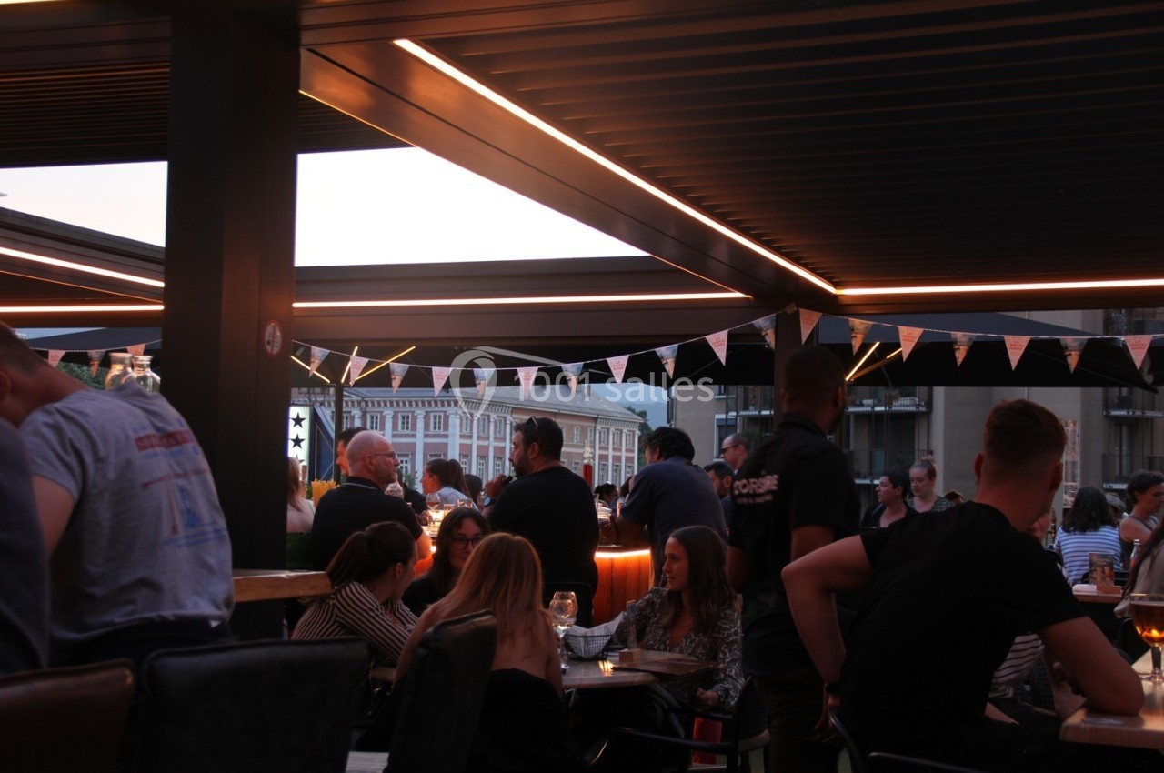 Terrasse animée en soirée avec des personnes discutant autour de tables, éclairage chaleureux et guirlandes décoratives.