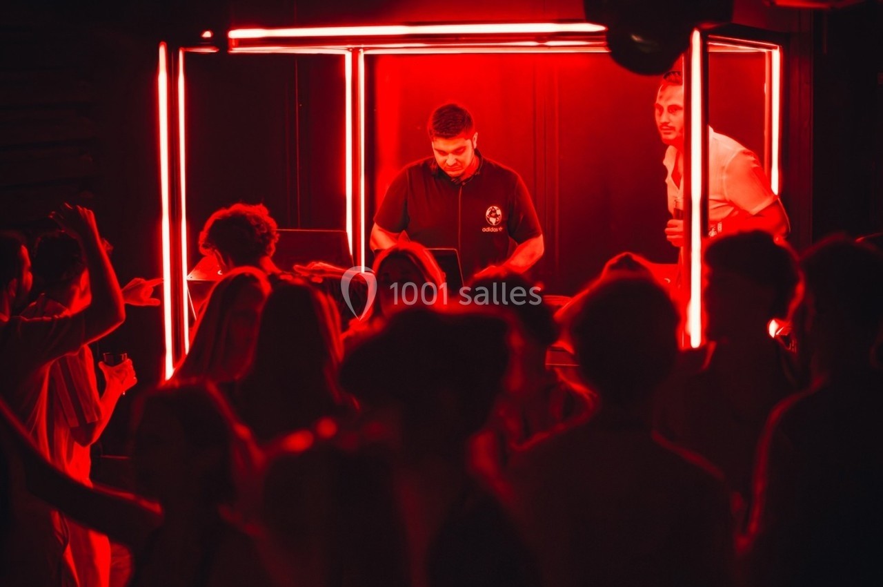 Un DJ mixe dans une cabine éclairée de rouge, entouré de personnes dansant dans une ambiance sombre.