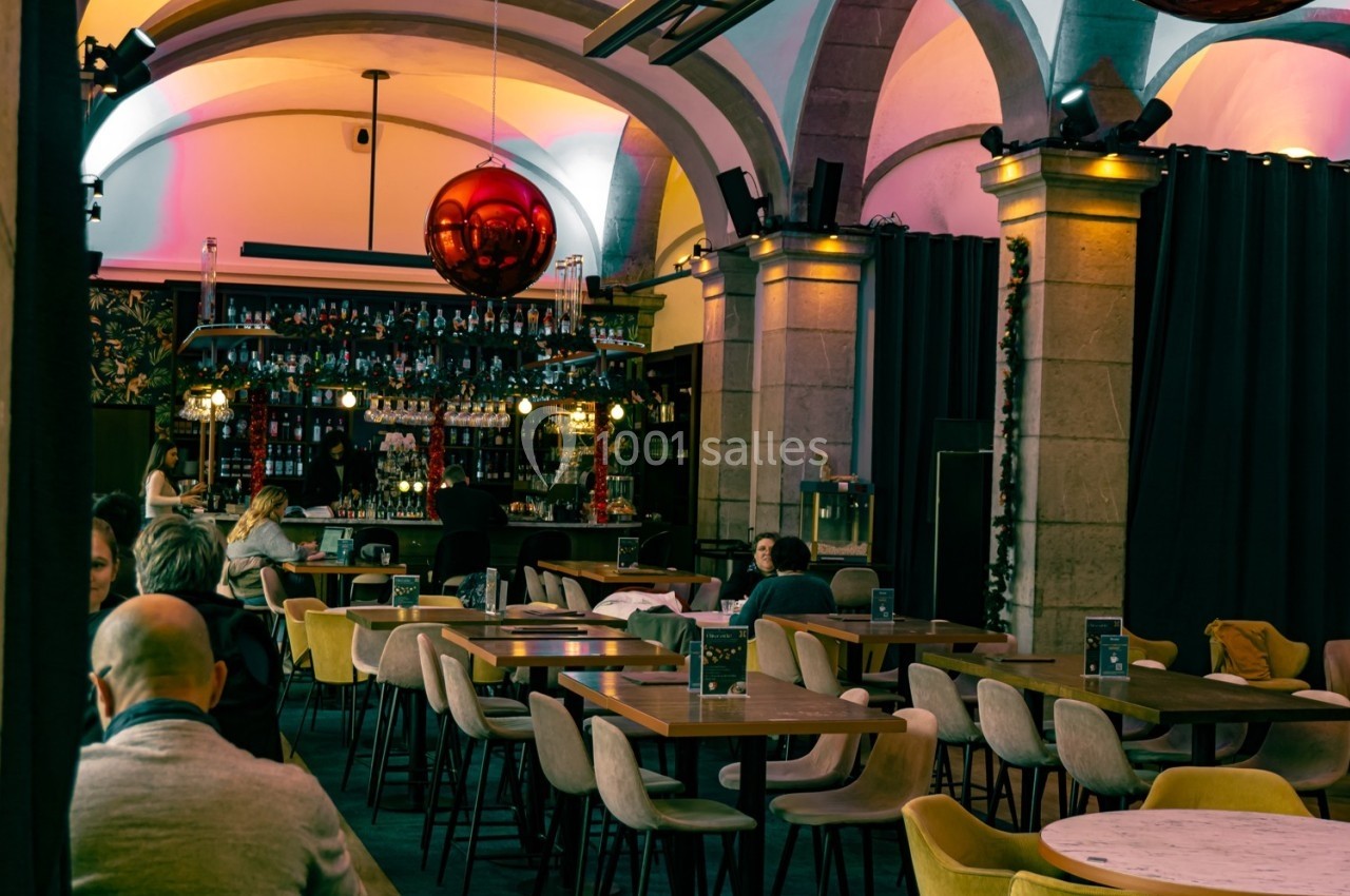 Salle de restaurant avec tables en bois, chaises rembourrées et un bar illuminé en arrière-plan.
