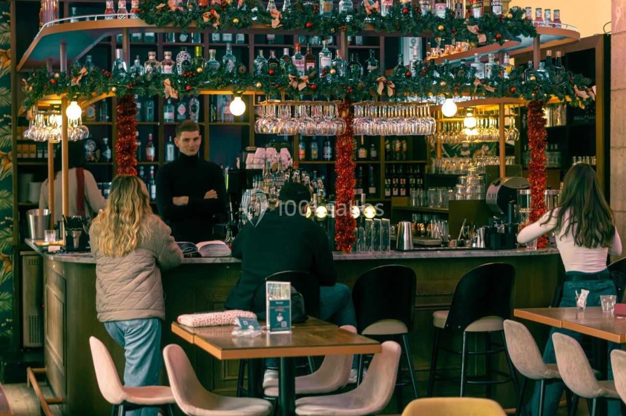 Bar décoré pour les fêtes avec des guirlandes, des clients assis au comptoir et une serveuse en arrière-plan.