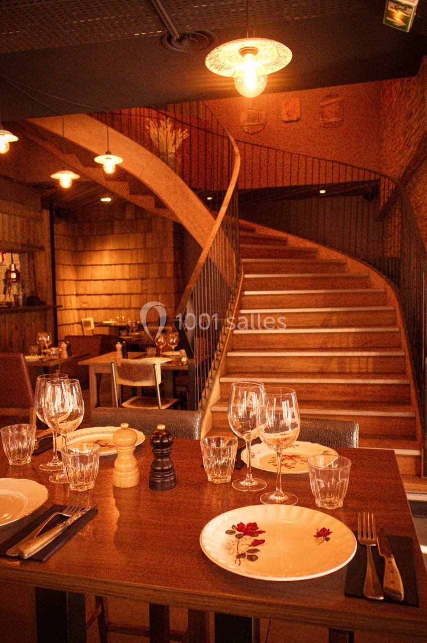 Table dressée avec vaisselle et verres dans un restaurant chaleureux, avec escalier en bois en arrière-plan.