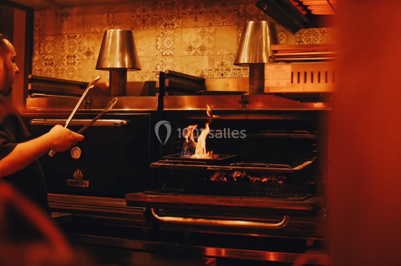 Un cuisinier manipule des pinces devant un four ouvert avec des flammes visibles sur un gril.