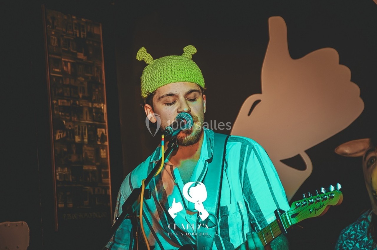 Un musicien portant un bonnet vert joue de la guitare et chante sur scène, avec un décor sombre en arrière-plan.