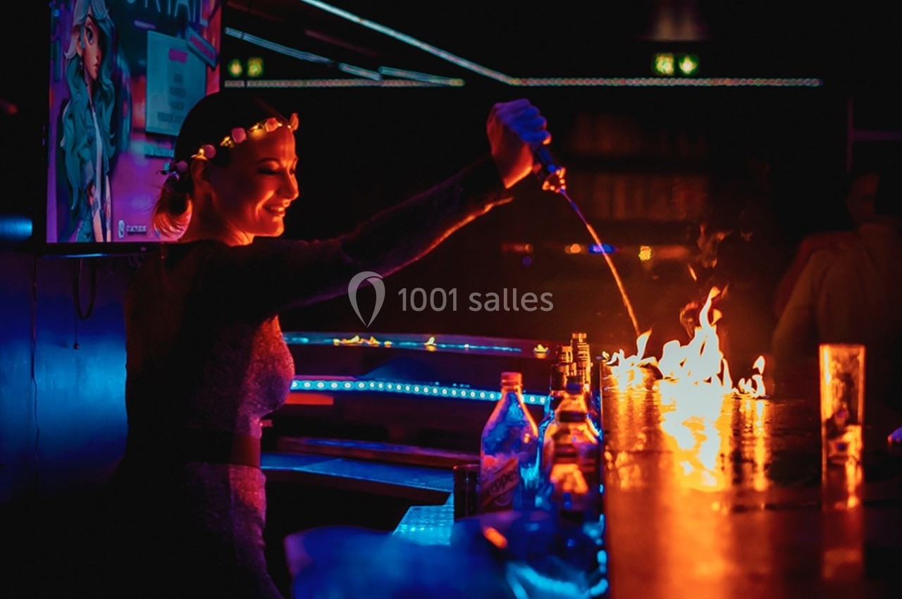 Une femme verse un liquide enflammé sur un bar dans une ambiance sombre et lumineuse de club.