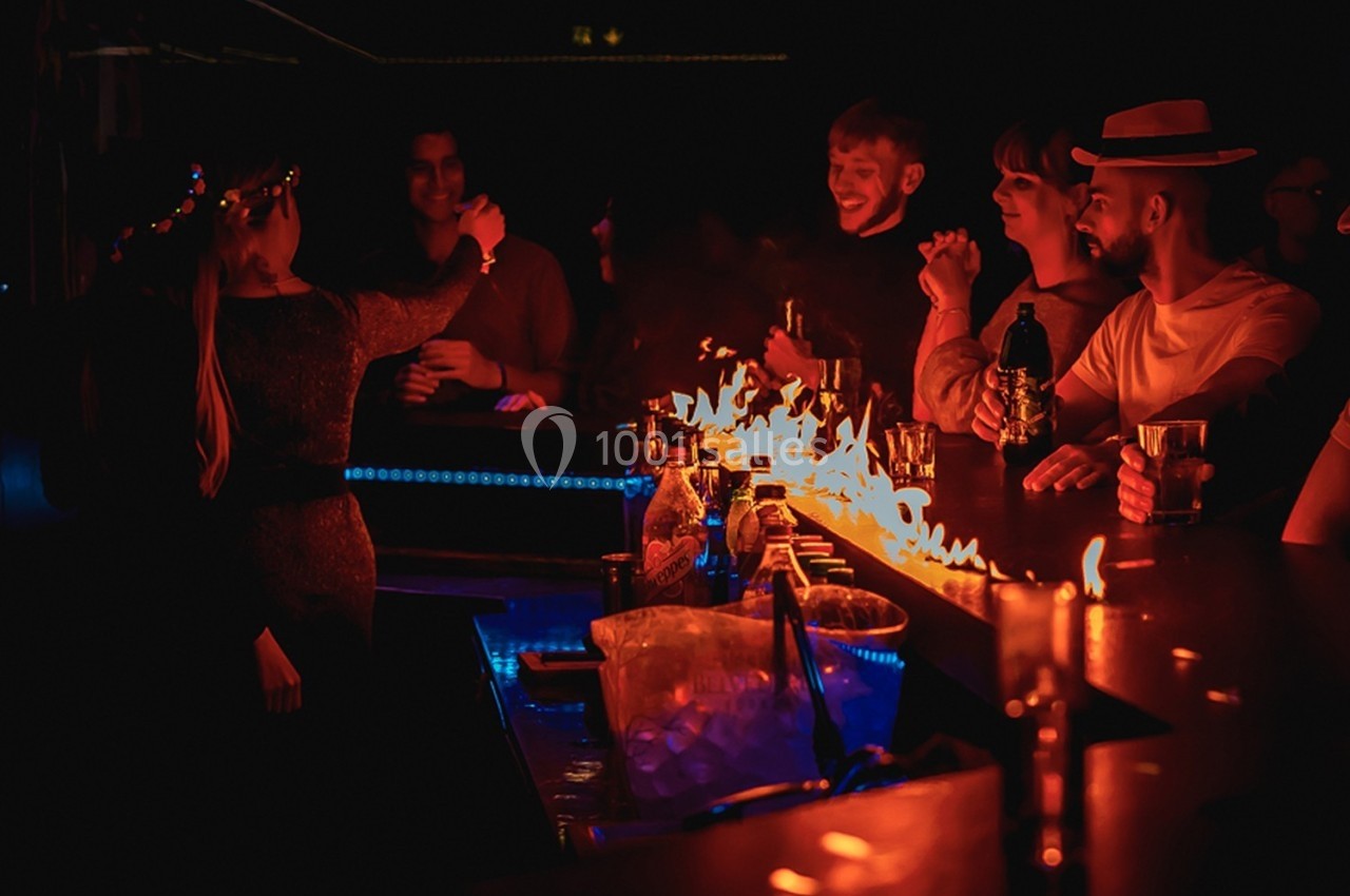 Des personnes discutent et boivent autour d'un bar illuminé par des flammes dans une ambiance sombre.