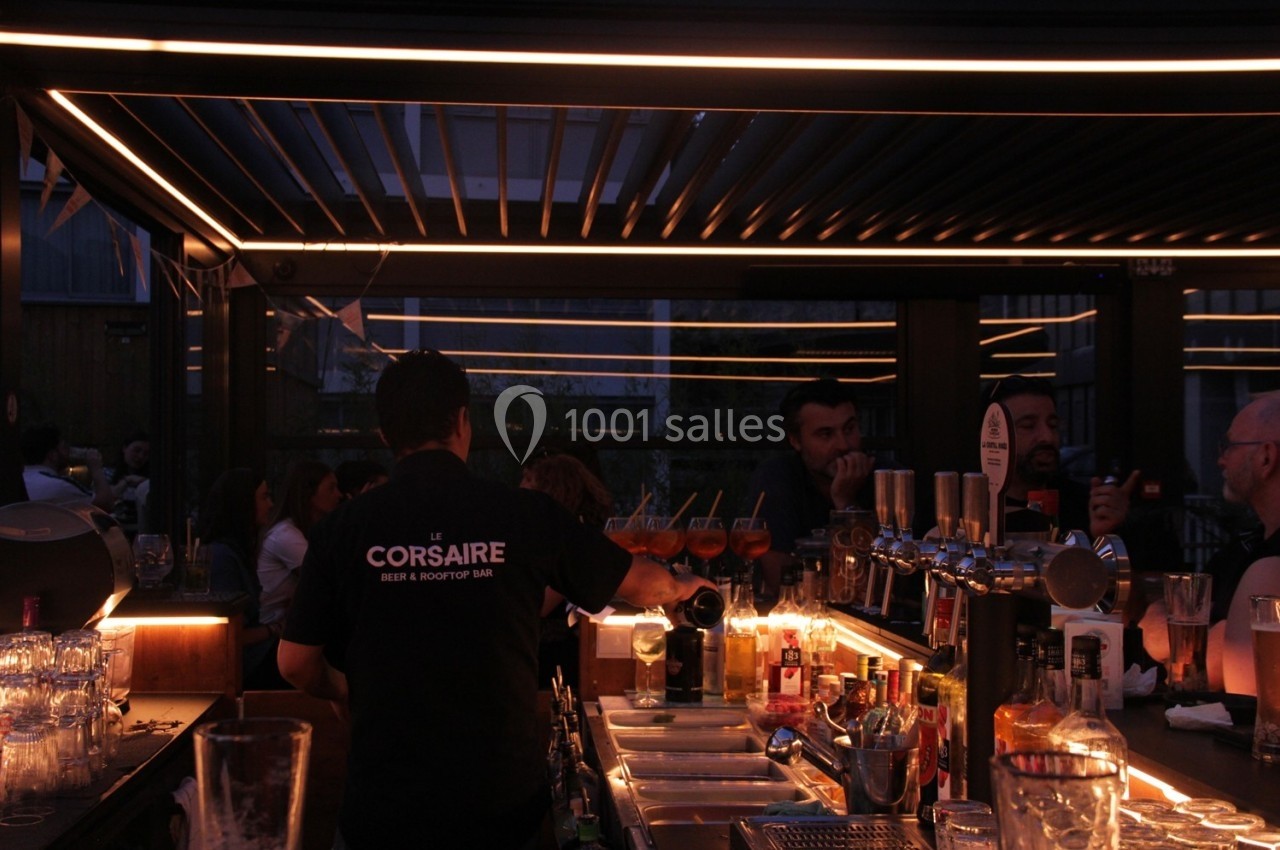 Un barman prépare des boissons dans un bar éclairé par des lumières tamisées, avec des clients en arrière-plan.