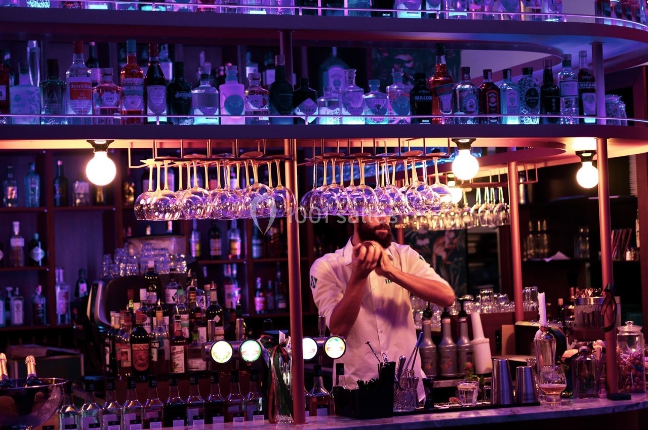 Un barman prépare un cocktail derrière un comptoir éclairé, entouré de bouteilles et de verres suspendus.