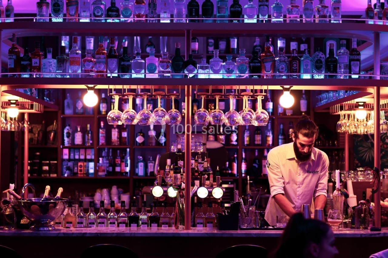 Un barman prépare des cocktails derrière un comptoir éclairé par des lumières violettes, entouré de bouteilles d'alcool.