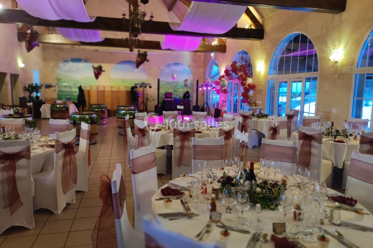 Salle de réception décorée pour un événement, avec tables dressées, chaises ornées de rubans et éclairage tamisé.
