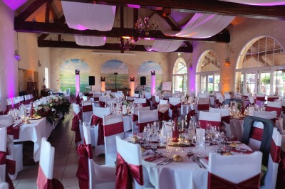 Salle de réception décorée avec des tables rondes dressées, nappes blanches et chaises ornées de nœuds rouges.