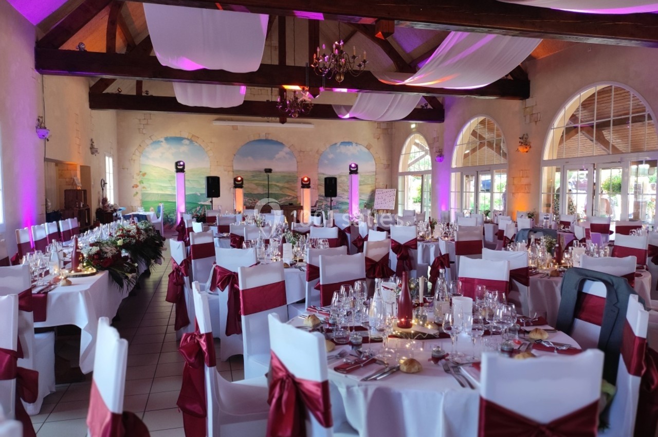 Salle de réception décorée pour un événement, avec des tables dressées, des chaises ornées de nœuds rouges et un éclairage…