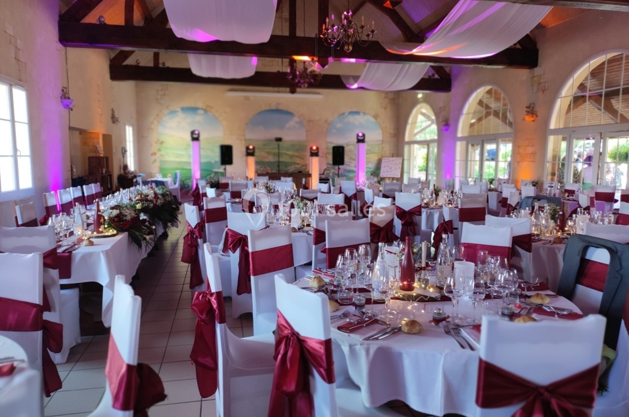 Salle de réception décorée pour un mariage avec nappes blanches, chaises ornées de rubans rouges et éclairage tamisé.