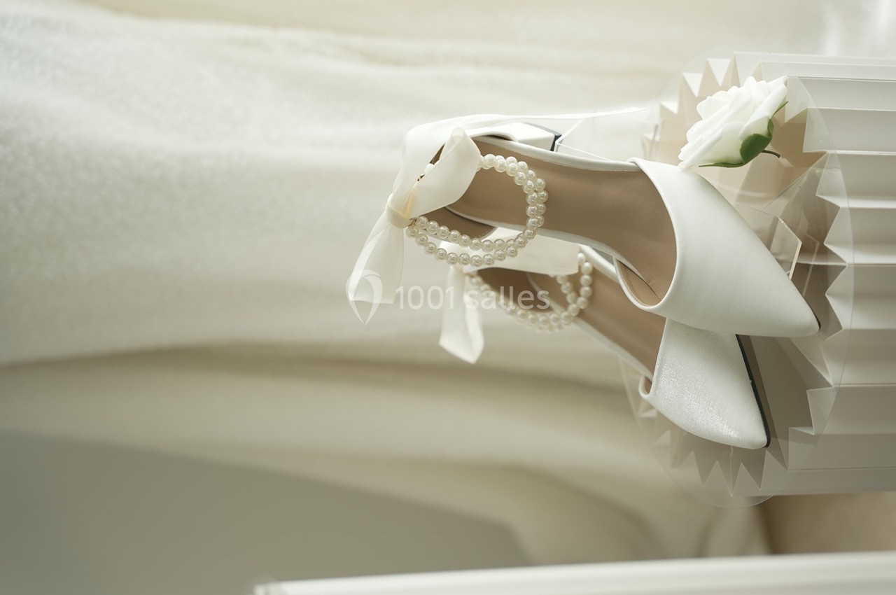 Chaussures blanches élégantes avec perles et ruban, posées sur une surface texturée près d'une fleur blanche.