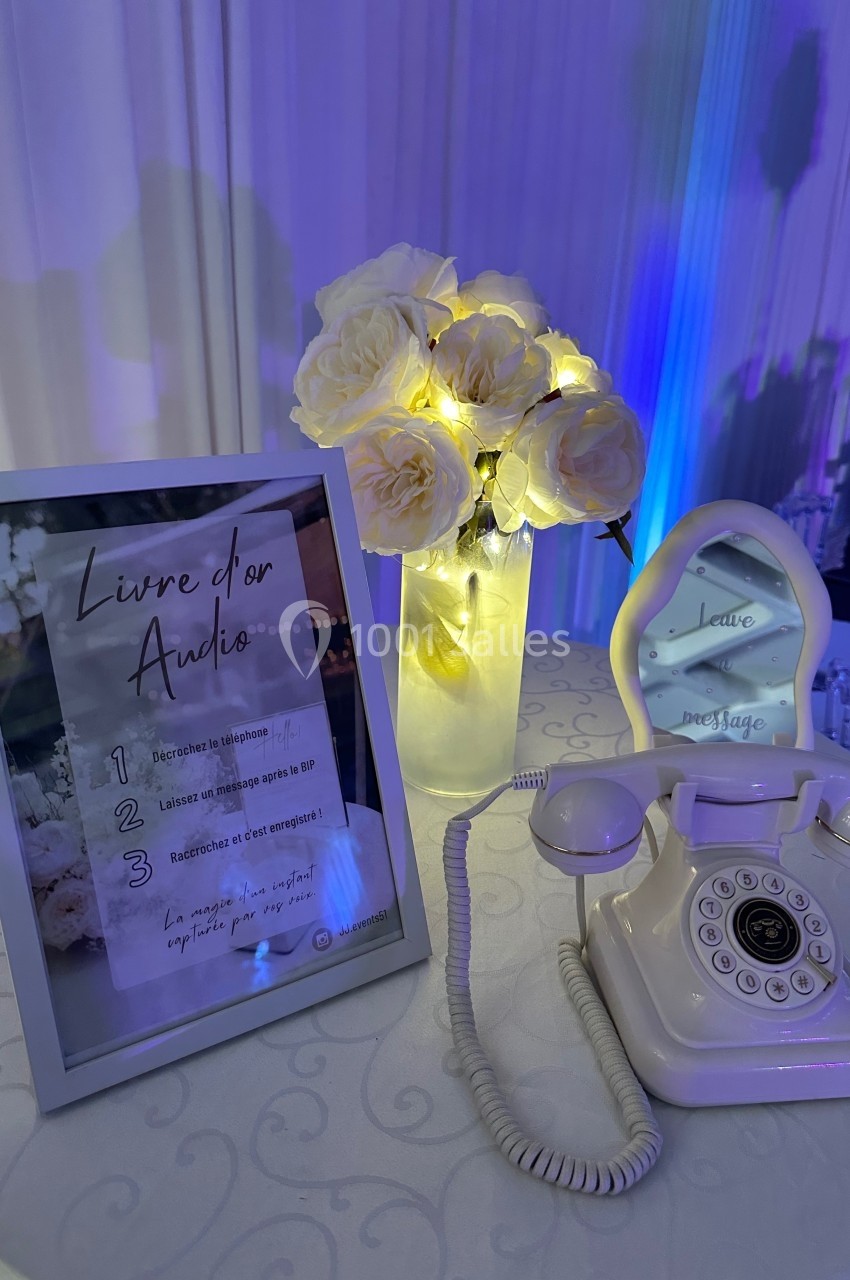 Un téléphone rétro blanc et un vase de roses illuminées sur une table, avec un cadre explicatif sur un livre d'or audio.