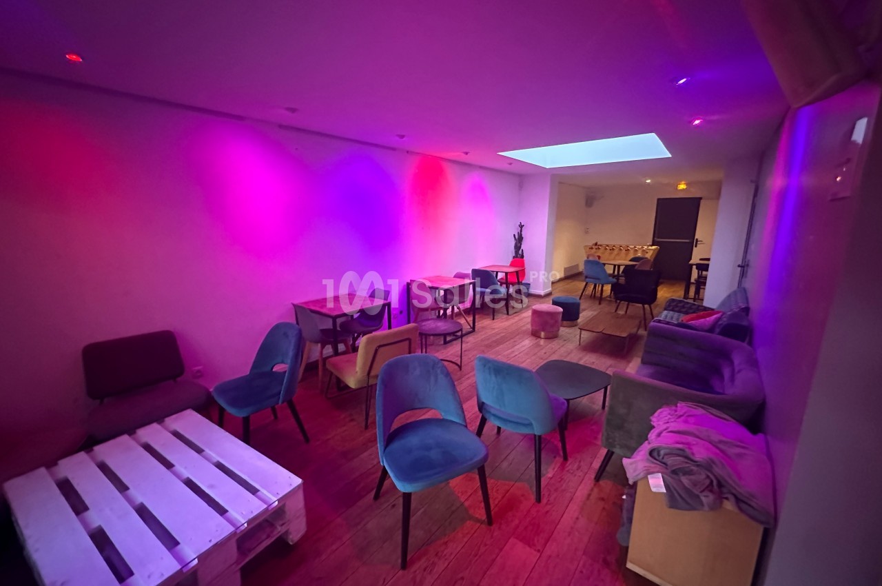 Salle avec éclairage tamisé coloré, mobilier varié comprenant chaises, tables et palettes en bois sur un sol en parquet.