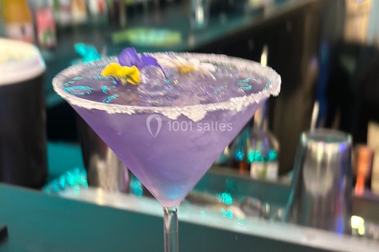 Cocktail violet servi dans un verre martini avec bord salé, décoré de fleurs comestibles, sur un bar.