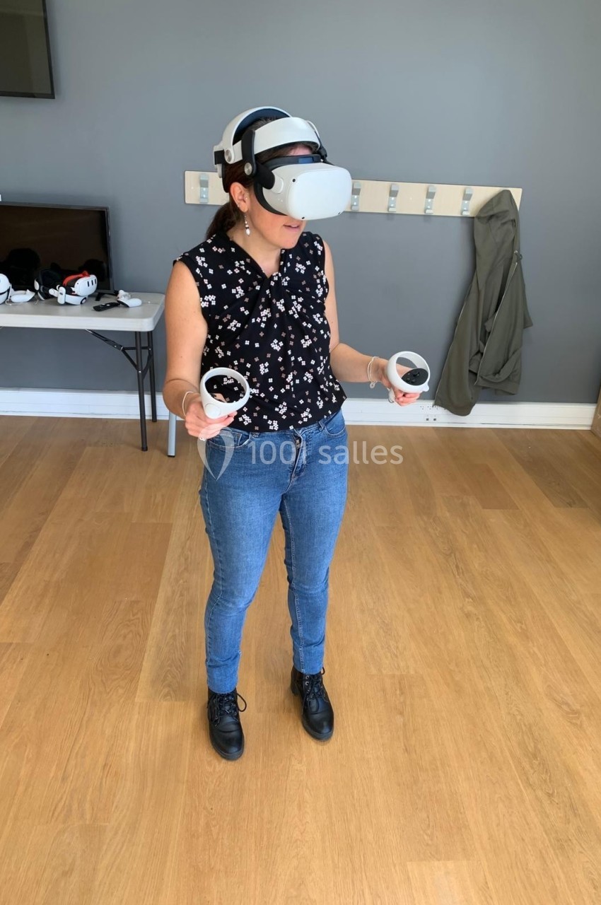 Une personne debout utilise un casque de réalité virtuelle avec des manettes, dans une pièce au sol en bois.