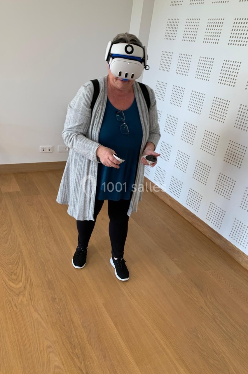 Femme portant un casque de réalité virtuelle, tenant des manettes, debout dans une pièce au sol en bois clair.