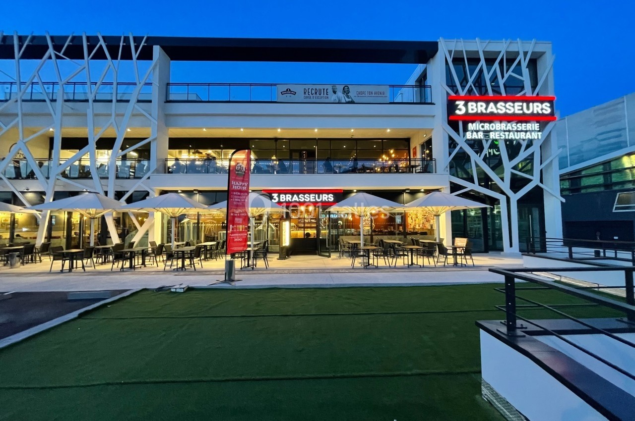 Façade éclairée d'un restaurant avec terrasse extérieure, enseignes lumineuses ’3 Brasseurs’, et ciel au crépuscule.