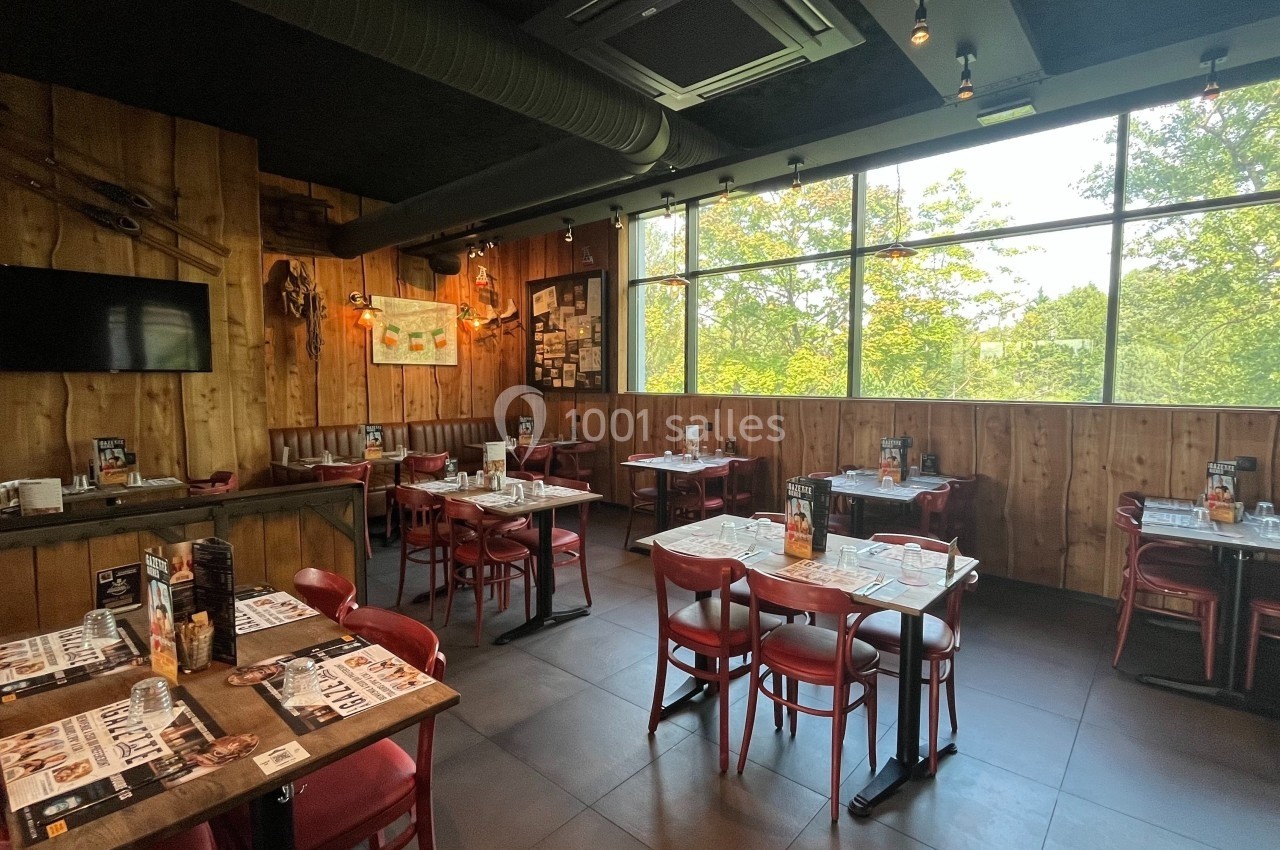Salle de restaurant lumineuse avec des tables en bois, des chaises rouges et une grande baie vitrée donnant sur des arbres.