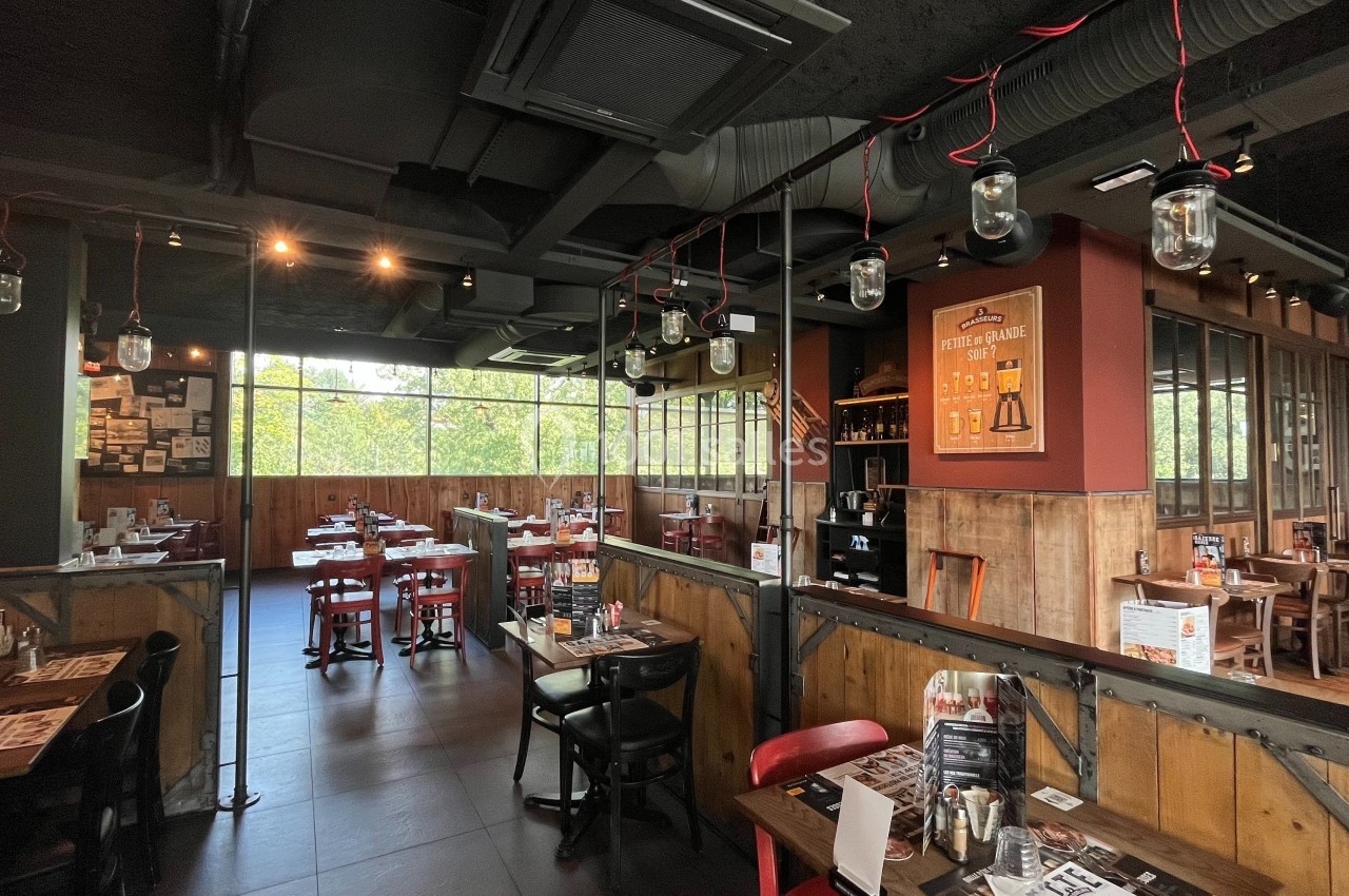 Salle de restaurant avec tables en bois, chaises rouges et noires, éclairage suspendu et grandes fenêtres donnant sur l…
