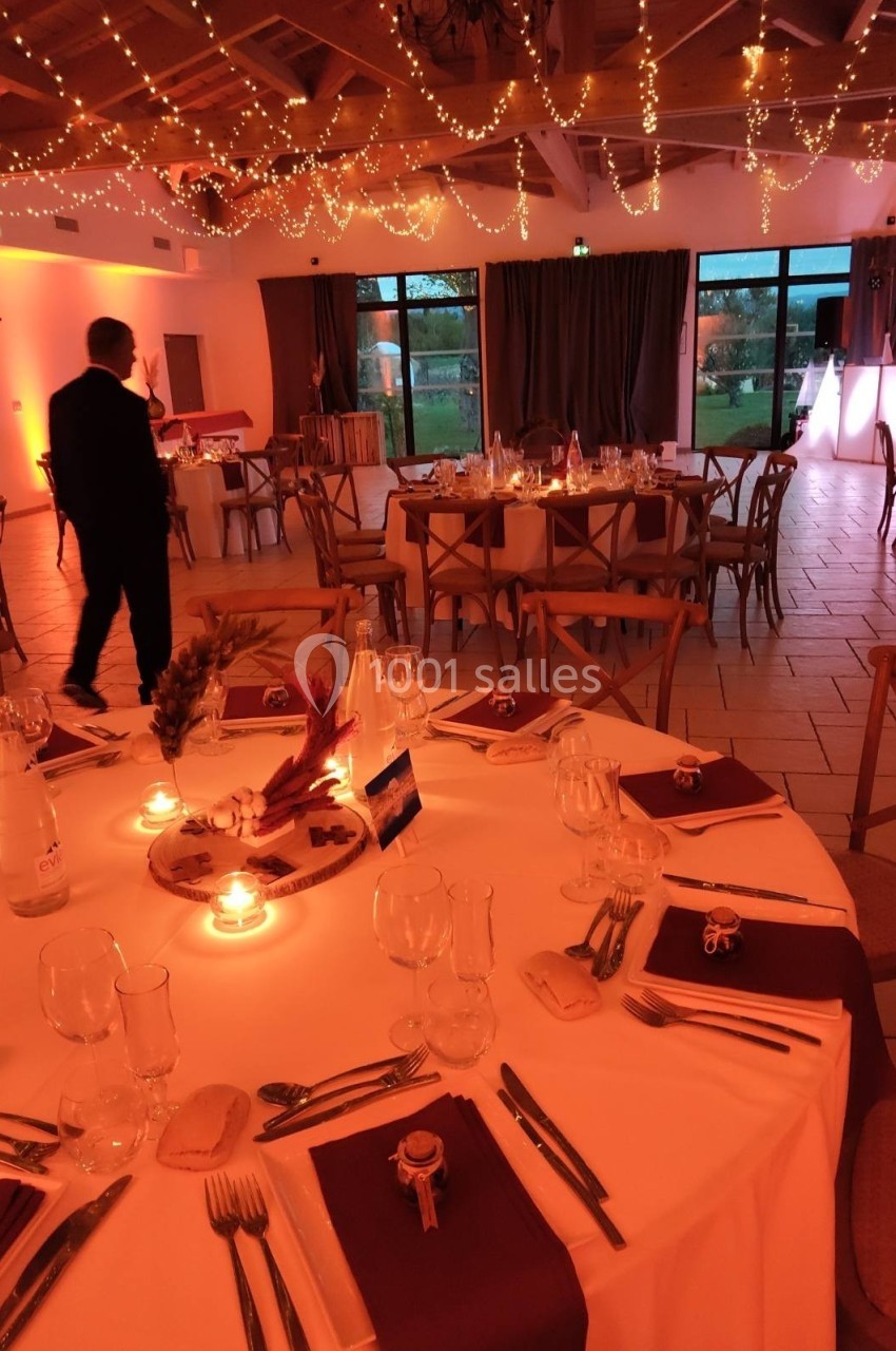 Salle de réception décorée avec des guirlandes lumineuses, tables dressées avec nappes blanches et serviettes bordeaux.