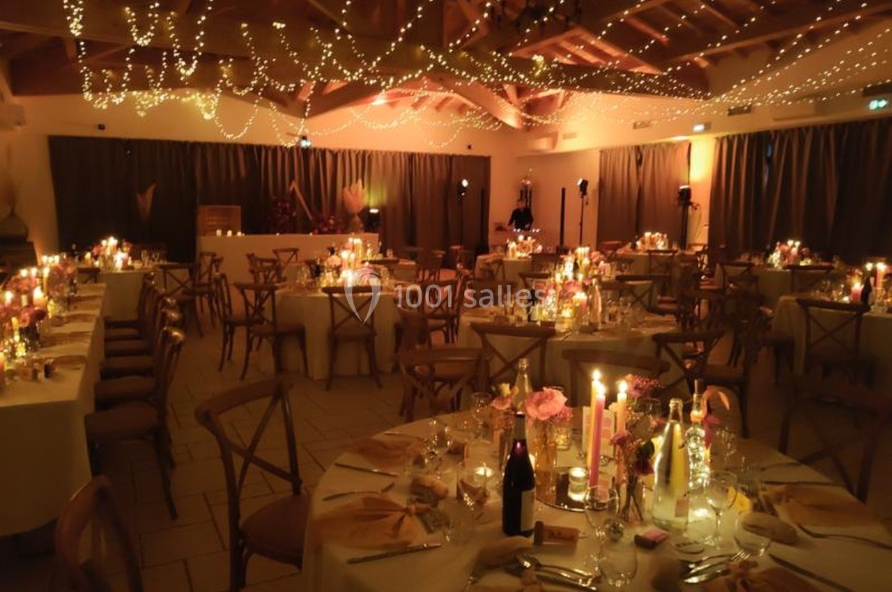 Salle de réception décorée pour un dîner, avec tables dressées, bougies allumées et guirlandes lumineuses suspendues.