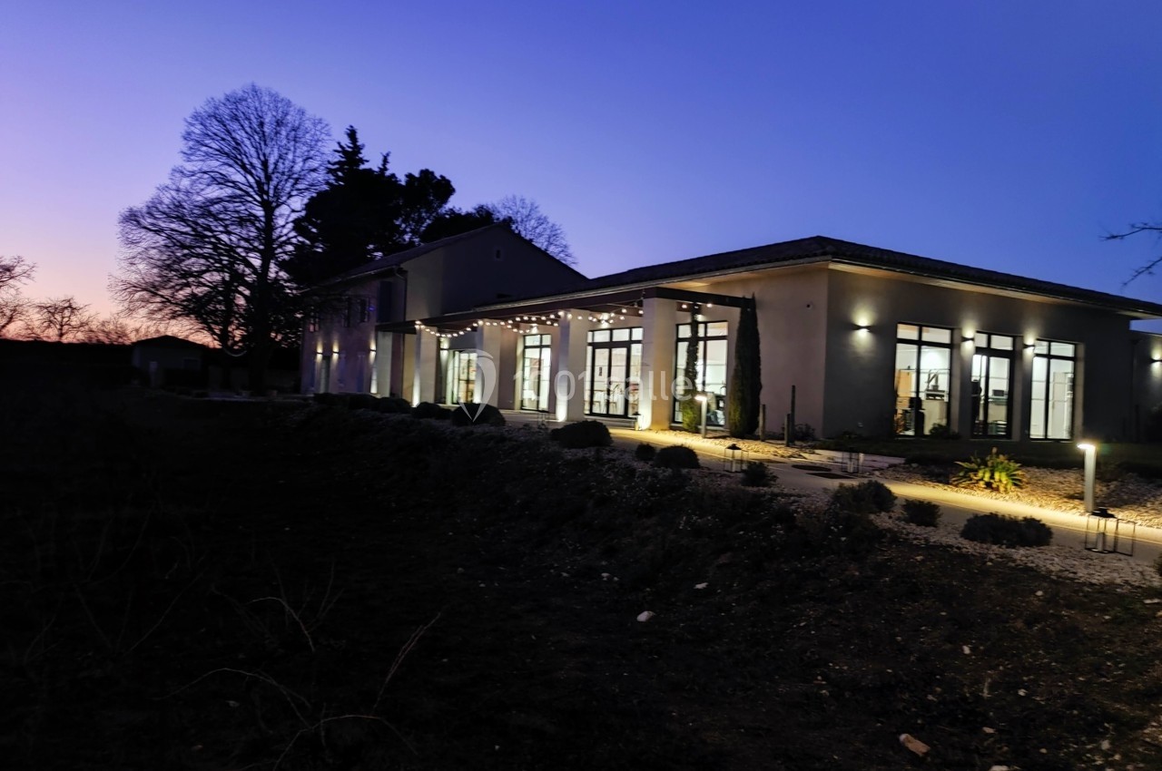 Bâtiment moderne éclairé en soirée, entouré d'arbres et d'un jardin sombre sous un ciel crépusculaire.