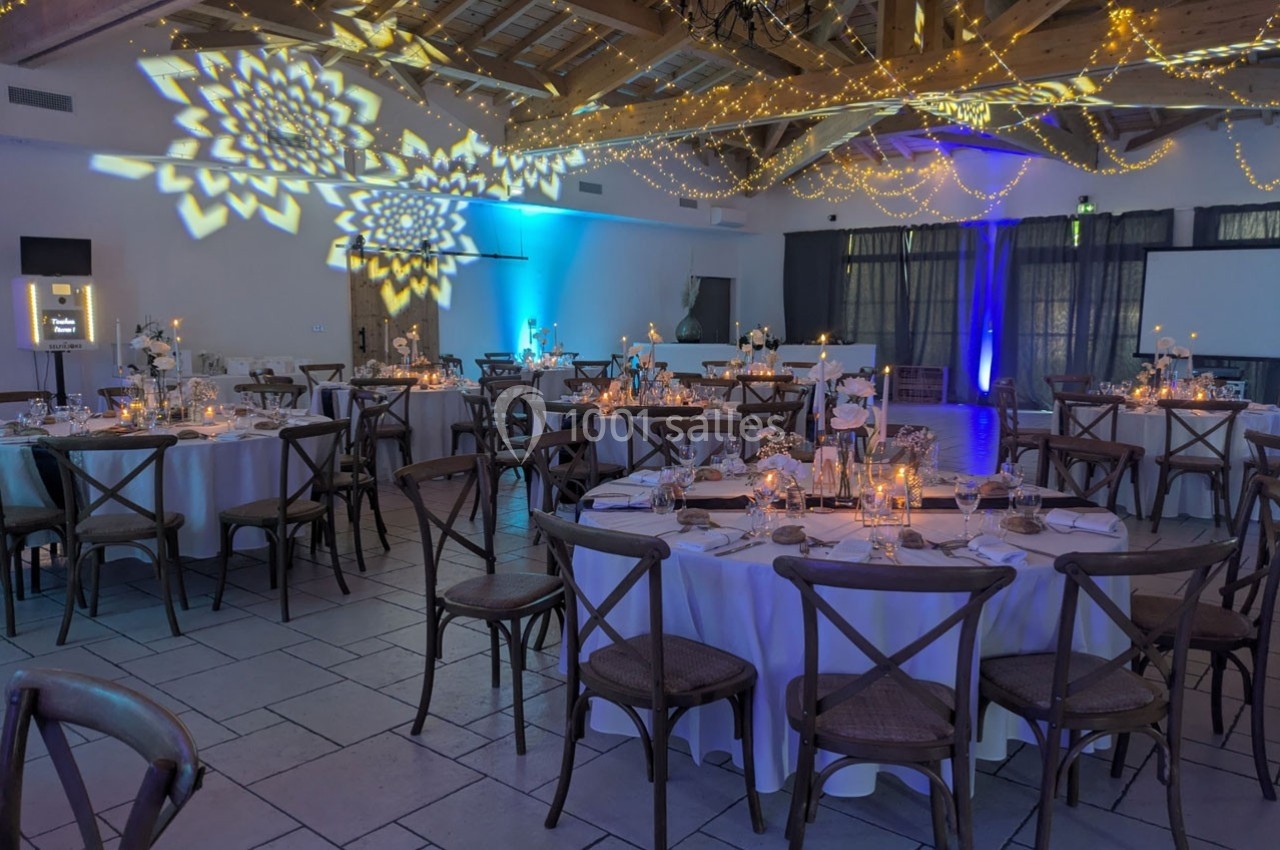 Salle de réception décorée avec des guirlandes lumineuses, tables rondes dressées et éclairage d'ambiance chaleureux.