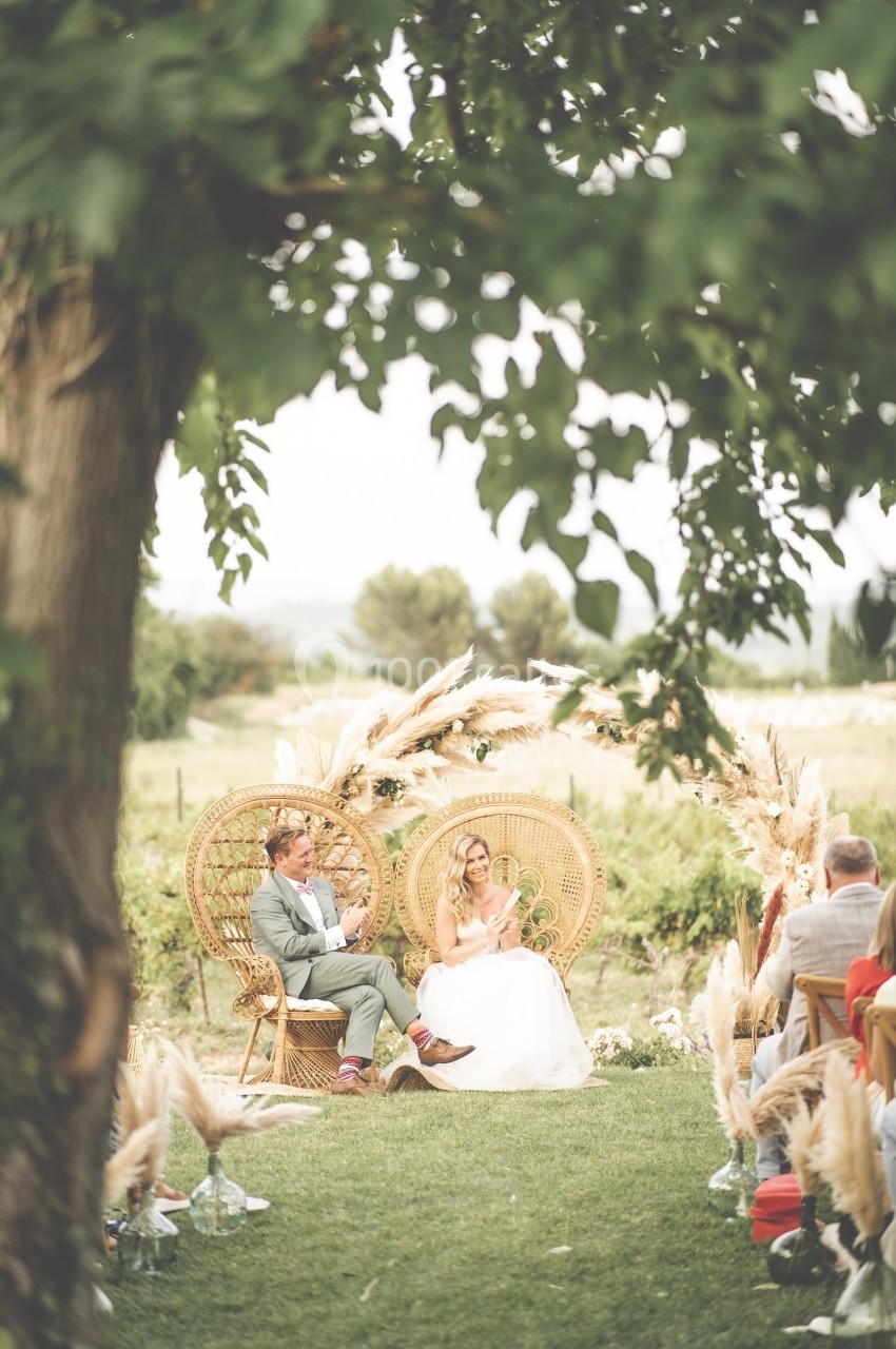 Un couple assis sur des fauteuils en rotin lors d'une cérémonie en extérieur, entouré de décorations végétales.