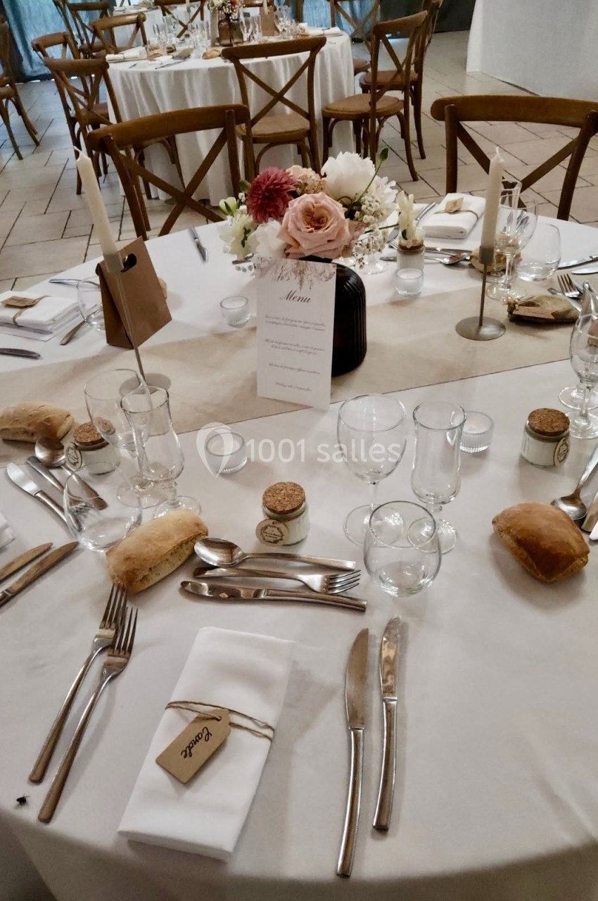 Table de réception décorée avec des fleurs, menus, couverts, verres et petits pains disposés sur une nappe blanche.