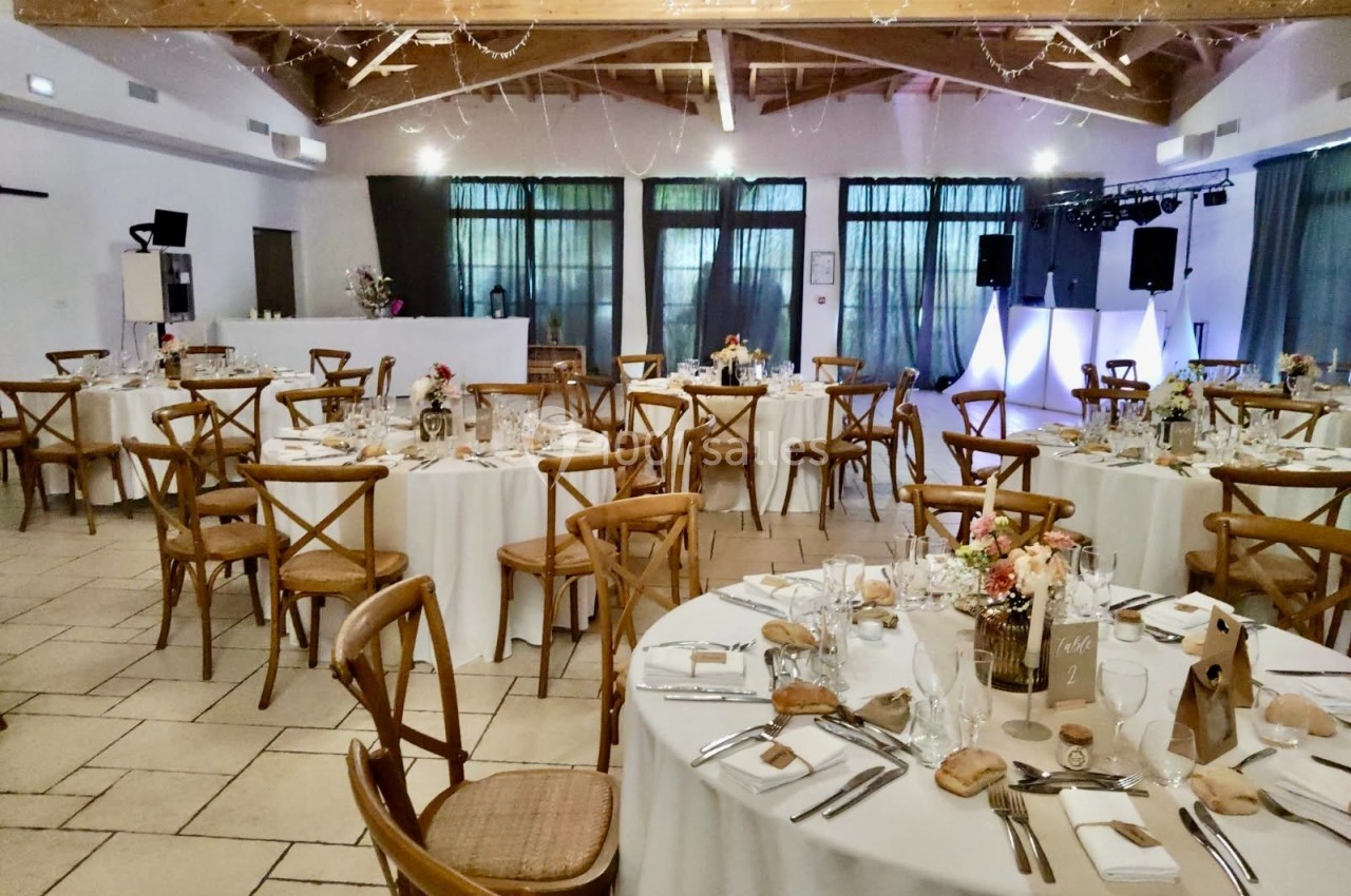 Salle de réception décorée avec des tables rondes dressées, chaises en bois et éclairage tamisé.
