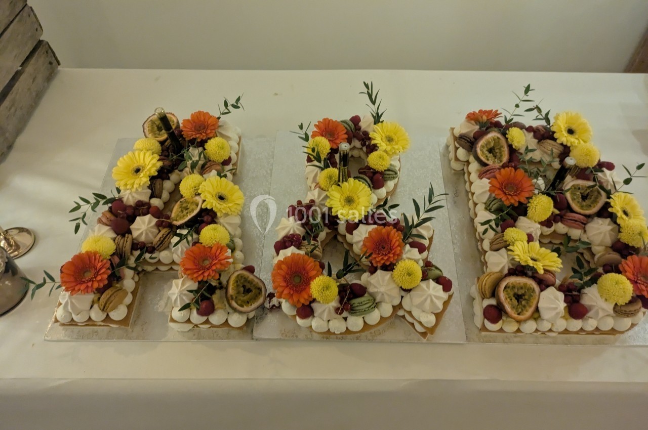 Gâteau en forme de lettres décoré de fleurs, fruits frais et meringues, posé sur une table blanche.